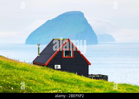 Schwarzes Haus auf dem berühmten färöischen Hexen Finger Trail und der Koltur Insel im Hintergrund. Dorf Sandavagur, Insel Vagar, Inseln der Färöer, Dänemark. Landschaftsfotografie Stockfoto