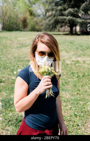 Junge Frau trägt chirurgische Maske versucht, wilde Blumen riechen Stockfoto