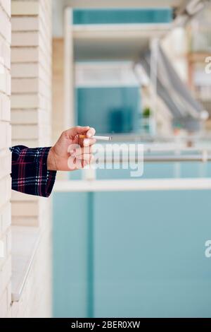 Nahaufnahme eines jungen kaukasischen Mannes, in Pyjamas, Rauchen einer Zigarette, die sich in der Fensterscheibe seines Hauses oder eines Hotels neigt Stockfoto