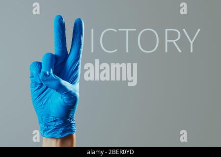 Nahaufnahme der Hand eines Mannes, mit blauen OP-Handschuhe, das V-Zeichen und den Rest der Buchstaben, um das Wort Sieg gegen eine graue Rückseite zu bilden Stockfoto