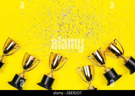 Goldene Siegerbecher mit silberfarbenen Glitzern auf gelbem Hintergrund mit Kopierraum. Festliches Konzept. Flacher Laienstil. Stockfoto
