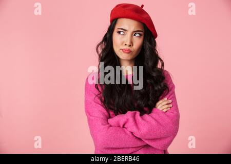 Bild der Unzufriedenheit schöne asiatische Mädchen mit langen dunklen Haaren tragen Baskenmütze stehend mit Armen gekreuzt isoliert über rosa Hintergrund Stockfoto