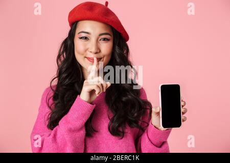 Bild von schönen asiatischen Mädchen tragen Baskenmütze halten Finger an den Lippen und zeigt Smartphone-Bildschirm isoliert über rosa Hintergrund Stockfoto