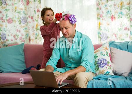 Kind spielt und stört Vater arbeitet fern von zu Hause. Kleines Mädchen Kämmen Papa's Haar und machen Frisur. Mann auf Couch mit Laptop Stockfoto