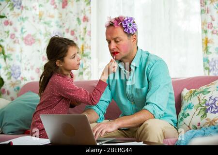 Kind spielt und stört Vater arbeitet fern von zu Hause. Kleines Mädchen, das Make-up anwendet. Mann auf der Couch mit Laptop. Familie Zeit zu verbringen Stockfoto