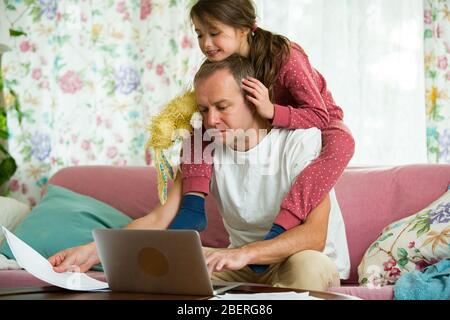 Kind spielt und stört Vater arbeitet fern von zu Hause. Mann auf der Couch mit Laptop. Familie verbringt Zeit zusammen drinnen. Stockfoto