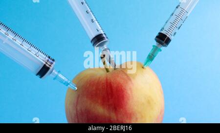 Drei verschiedene medizinische Spritzen, die tief in einen Apfel auf blauem Hintergrund stecken. Die Frucht ist mit Spritzen übersät. Ein reifer roter Apfel, der von al durchbohrt wurde Stockfoto