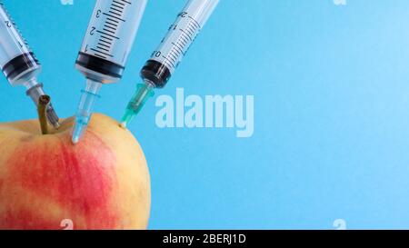 Drei verschiedene medizinische Spritzen, die tief in einen Apfel auf blauem Hintergrund stecken. Die Frucht ist mit Spritzen übersät. Ein reifer roter Apfel, der von al durchbohrt wurde Stockfoto
