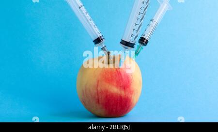 Drei verschiedene medizinische Spritzen, die tief in einen Apfel auf blauem Hintergrund stecken. Die Frucht ist mit Spritzen übersät. Ein reifer roter Apfel, der von al durchbohrt wurde Stockfoto
