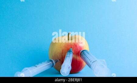 Drei verschiedene medizinische Spritzen, die tief in einen Apfel auf blauem Hintergrund stecken. Die Frucht ist mit Spritzen übersät. Ein reifer roter Apfel, der von al durchbohrt wurde Stockfoto