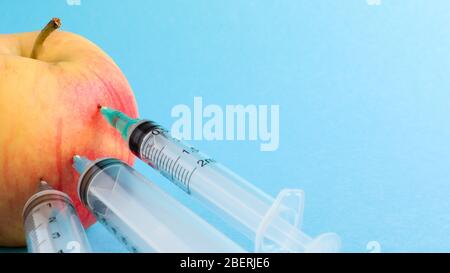 Drei verschiedene medizinische Spritzen, die tief in einen Apfel auf blauem Hintergrund stecken. Die Frucht ist mit Spritzen übersät. Ein reifer roter Apfel, der von al durchbohrt wurde Stockfoto