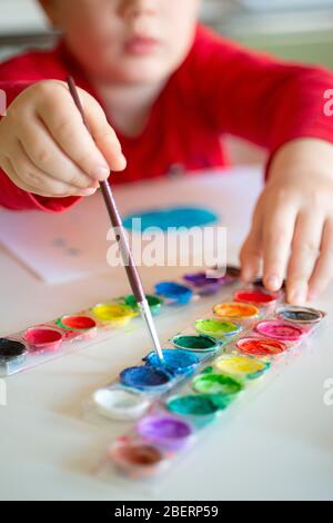 Der Junge lernt, mit einem Pinsel auf Papier zu malen, der an einem Tisch sitzt Stockfoto