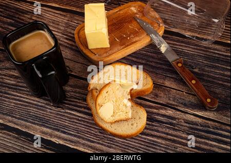 Frischer Weizenstoast mit Butter, ein keramischer Becher Kaffee mit Milch und ein Stück Butter in einer hölzernen Butterdose auf einem hölzernen Hintergrund. Nahaufnahme Stockfoto