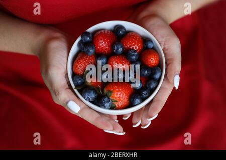 Mädchen in rotem Kleid hält Teller mit Beeren. Weißer Teller mit Erdbeere und Heidelbeere. Gesunde Lebensweise. Fruchtvitamin. Obstsalat Stockfoto