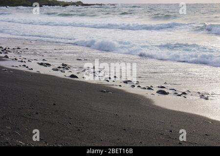 Kleine Wellen Rollen auf einen schwarzen Sandstrand mit Felsen und Kieselsteinen in Hana Bay, Maui, Hawaii, USA Stockfoto