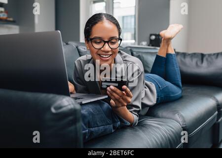 Frau in Brille, die zu Hause auf einem Sofa liegt und eine Kreditkarte hält. Lächelnde Frau, die eine Online-Zahlung auf ihrem Laptop-Computer macht. Stockfoto