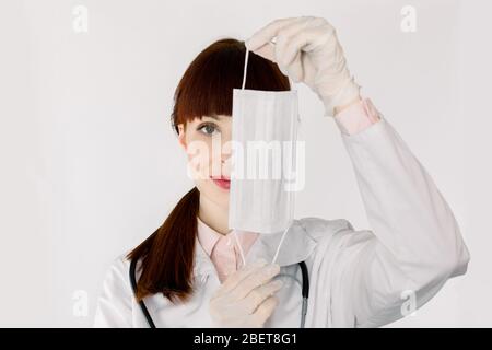 Junge ziemlich dunkelhaarige Frau Arzt oder Krankenschwester, trägt weißen Mantel, versteckt ihr Gesicht hinter einer medizinischen Maske, posiert auf isolierten weißen Hintergrund, Kopie Stockfoto