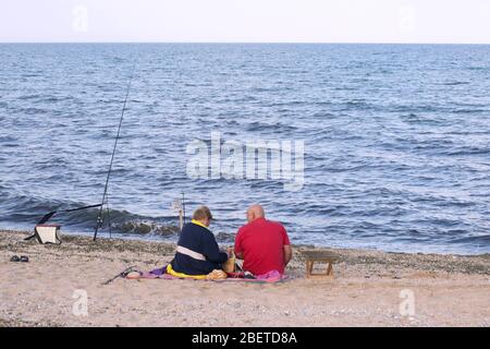 Seniorenpaar sitzt am Strand auf einem Picknick mit Angelruten am Meer Stockfoto