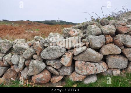 Flechten bedeckte Granitfelsen bauten auf der Insel St. Agnes auf den Inseln Scilly eine Steinmauer mit dem alten Leuchtturm im Hintergrund. Stockfoto