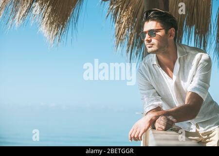 Junger attraktiver Mann mit Sonnenbrille, der im Sommer über das Meer blickt. Er blickt nach vorne, in ein weißes Hemd gekleidet und lehnt sich auf ein Holz Stockfoto