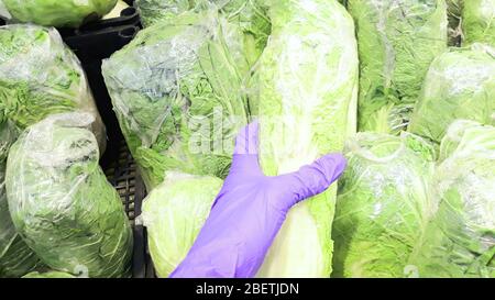 Hand in einem blauen medizinischen Handschuh hält grünen Pekingkohl in Lebensmittel Cellophan in einem Quarantänestore während der Coronavirus-Epidemie verpackt, COVID-19 Stockfoto