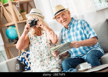Ältere Paar sitzt in Reisebüro sitzen mit Hüten halten Bilder und halten Karte Blick Kamera fröhlich Stockfoto