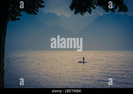 Blick auf die Alpen, Figur Silhouette eines Mannes auf einem Surfbrett mit Paddel auf dem Genfer See in Montreux, Schweizer Riviera, Kanton Waadt, Stockfoto