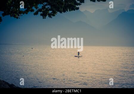 Blick auf die Alpen, Figur Silhouette eines Mannes auf einem Surfbrett mit Paddel auf dem Genfer See in Montreux, Schweizer Riviera, Kanton Waadt, Stockfoto