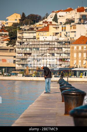 Älterer Mann, der von einem Weg aus gesehen wird, der einen Hafenpier hinunter geht und auf das Meer blickt. Gebäude in Split Küste in der Ferne Stockfoto
