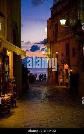 Nächtliche Straßen mit hellen Laternen Lampen auf Gebäuden mit Balkonen, Silhouette von Menschen und bunten Himmel mit Wolken auf dem Hintergrund, Tropea Stockfoto