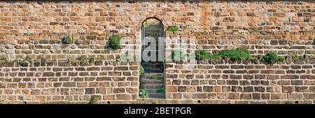 Alte gelbe und braune Sandsteinwand, Stufen (Treppe) aus Blöcken von unregelmäßiger Form Hintergrund und Metall-Tor Stockfoto