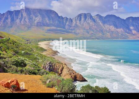 Wunderschöne südafrikanische Küste entlang der Garden Route mit einer malerischen Bucht mit einem langen, einsamen Strand und hohen Bergen im Hintergrund. Stockfoto