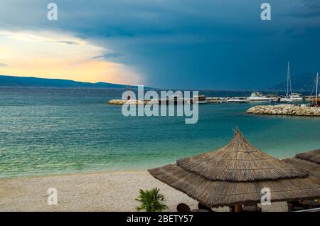 Schöne Aussicht auf Sandstrand mit Strohschirmen, Hafen und kleinen Leuchtturm auf Steinpier vor der Insel Brac am Vorabend des Sturmregens, Baska Vo Stockfoto