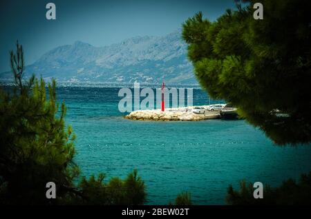 Schöne Aussicht auf kleinen Leuchtturm auf Steinsteg durch grüne Nadelbäume vor Biokovo Bergkette, Baska Voda, Makarska riviera, Dalm Stockfoto