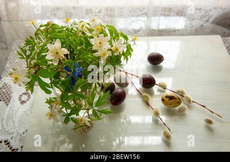 Weißer Wald Frühlingsblumen in einem Glas, Ostereier Wachtel und Weidenzweige auf dem Hintergrund von Spitzen Vorhänge. Stockfoto