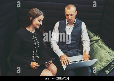 Eine elegante reife Geschäftsfrau mit einem digitalen Tablet in den Händen schaut auf dem Bildschirm des Laptops ihres männlichen Kollegen neben ihr sitzen Stockfoto