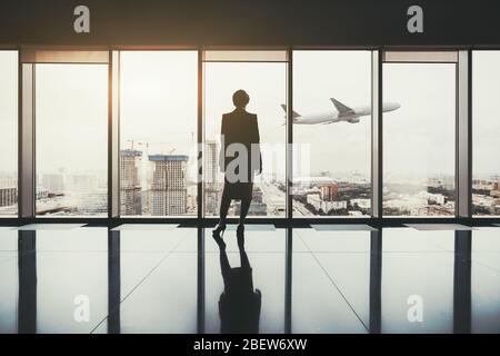 Eine Silhouette einer erfolgreichen Unternehmerin, die drinnen von einem luxuriösen Interieur mit einem reflektierenden Marmorboden steht, und einem Flugzeug, das an Höhe gewinnt Stockfoto