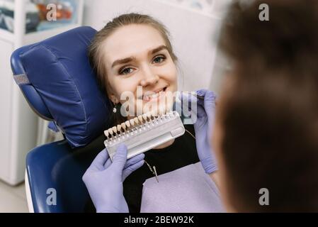 Zahnarzt in blauen medizinischen Handschuhen Anwendung Probe von Zahnschmelz Skala zu glücklichen Frau Patienten Zähne, um den richtigen Schatten, Zähne Bleichen Verfahren, Stockfoto