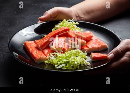 Krabbenstäbchen mit grünem frischen сhinese-Kohl. Weibliche Köchin Hände mit einem schwarzen Teller. Imitierte Meeresfrüchte. Zutaten für Salatmischung Stockfoto