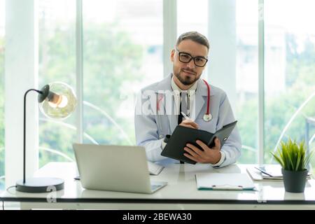 Doktor Mann sitzt am Schreibtisch an seinem Arbeitsplatz und lächelt in die Kamera. Perfekter medizinischer Service in der Klinik. Glückliche Zukunft der Medizin und des Gesundheitswesens. Stockfoto