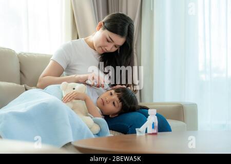 Asiatische junge Mutter Überprüfung der Messtemperatur ihrer kranken kleinen Tochter Gefühl wie Fieber oder Grippe auf dem Sofa zu Hause. Bilder von Menschen, die sich krank fühlen Stockfoto