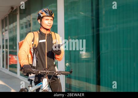 Asiatische Mann Kurier Überprüfung Kundenadresse in der Karte in Smartphone auf Fahrrad liefern Lebensmittel in der Stadt Straßen mit einem warmen Essen Lieferung von Take Aways A Stockfoto