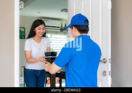 Asiatische Lieferung junger Mann in blauem, gleichmäßigem Lächeln und mit Essenskisten im Vorderhaus und Asian Woman, die eine Lieferung von Lebensmittelkisten aus Deliverym annehmen Stockfoto