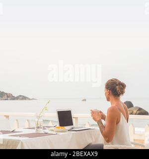 Eine Frau, die mit ihrem Laptop am frühen Morgen in einem Restaurant am Strand sitzt und Kaffee trinkt. Stockfoto