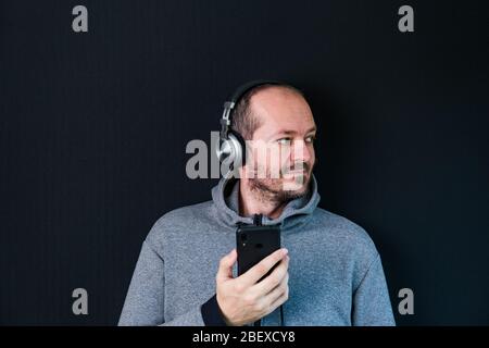 Bärtiger entspannter Mann im Hoodie, der vor schwarzem Hintergrund mit Kopfhörern steht, Musik hört und lächelnd Kopf und Schultern Stockfoto