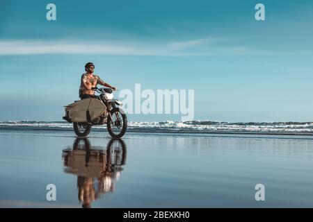 Echte Bali Male Surfer fährt auf Motorrad mit Surfbrett am Meeresstrand Stockfoto