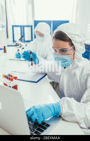 Biotechnologe, der das Reagenzglas mit Blutprobe hält und einen Laptop benutzt Stockfoto