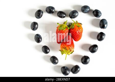Schwarze Trauben in Form eines Liebesherzens mit drei Erdbeeren in der Mitte vor einem schlichten weißen Hintergrund Stockfoto