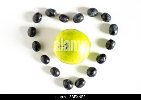 Schwarze Trauben in Form eines Liebesherzens mit einem Granny smith Apfel in der Mitte vor einem schlichten weißen Hintergrund ausgelegt Stockfoto