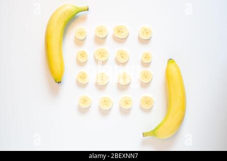 Eine Reihe von in Scheiben geschnittenen Bananenstücken, die in einem quadratischen Muster mit zwei vollen Bananen entweder Ende vor einem schlichten weißen Hintergrund ausgelegt sind Stockfoto
